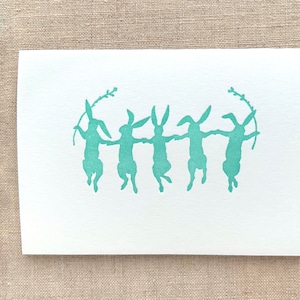 May include: A white greeting card with a teal illustration of five rabbits holding hands in a circle. Each rabbit holds a sprig. A sprig of pussy willow is in the upper left corner.