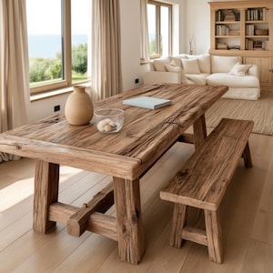 Op de afbeelding: Een rechthoekige eettafel en een bijpassende bank gemaakt van verweerd, lichtbruin hout met een zichtbare nerf. Op de tafel staan een gladde, beige keramische vaas, een doorzichtige glazen kom met kleine witte steentjes en een lichtblauw boek.