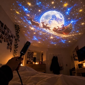May include: A black handheld projector device casting a vibrant light display onto a ceiling, featuring a large full moon, a silhouette of Santa Claus in a sleigh pulled by reindeer, and a field of glowing yellow stars against a blue nebula.