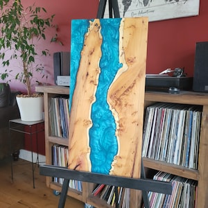 May include: A rectangular wooden slab featuring a central river-like inlay of vibrant, shimmering turquoise epoxy resin. The natural wood grain displays light tan tones with visible knots and organic, irregular edges along the resin channel.