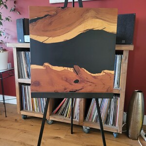 May include: A square wooden panel featuring a natural live edge grain pattern separated by a wide, smooth, matte black resin inlay that runs horizontally across the center, displayed on a black metal tripod easel stand.