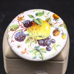 Op de afbeelding: Een witte keramische sieradenbox met een rond deksel met een kleurrijk fruit- en bloemendesign. Het deksel heeft een gele appel, rode en zwarte bessen en groene bladeren op een witte achtergrond.
