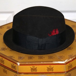 Op de afbeelding: Een zwarte fedora hoed met een zwarte band en een rode veersiering. De hoed ligt op een vintage zeshoekige hoedendoos met een goud-bruin ontwerp. De hoedendoos heeft een decoratieve rand.