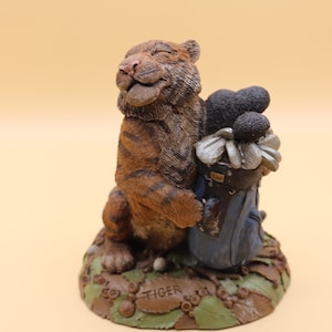 May include: A ceramic figurine of a tiger holding a golf bag with golf clubs. The tiger is smiling and has its eyes closed. The base of the figurine is decorated with green leaves and brown acorns. The base is also inscribed with the word "TIGER".