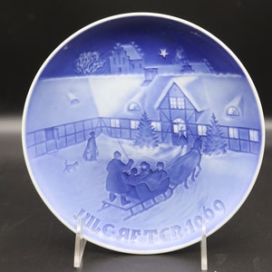 Könnte beinhalten: Ein blauer und weißer Weihnachtsteller mit einer Winterszene. Der Teller zeigt ein Haus mit einem schneebedeckten Dach, eine Gruppe von Menschen, die in einem Schlitten fahren, und einen Stern am Himmel. Der Text "JULGAFTER 1969" ist auf der Unterseite des Tellers gedruckt.