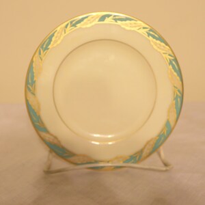 May include: A small, white ceramic plate with a green and gold rim. The rim features a repeating pattern of stylized leaves.