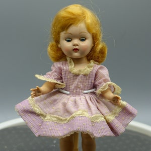 Könnte beinhalten: Eine Vintage-Puppe mit blonden Haaren, die ein rosa-weißes Gingham-Kleid mit Spitzenbesatz trägt. Die Puppe steht auf einem braunen Sockel.