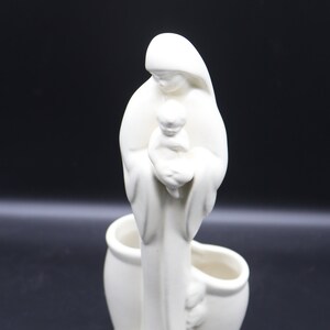 Op de afbeelding: Witte keramische plantenbak met een gesculpteerd beeld van een vrouw die een kind vasthoudt. De plantenbak heeft twee aparte compartimenten voor planten.