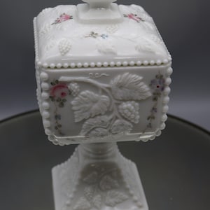 May include: White milk glass candy dish with a square base and lid. The dish is decorated with a raised design of grapes and leaves. The lid has a knob in the center and is also decorated with grapes and leaves. The dish is hand-painted with pink and blue flowers.