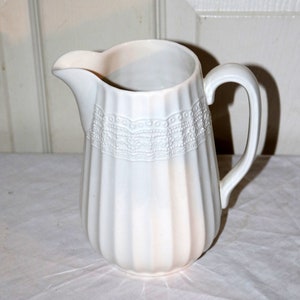 May include: A white ceramic pitcher with a ribbed body and a decorative lace-like band near the top. The pitcher has a curved handle and a spout for pouring. The pitcher is likely used for serving beverages.