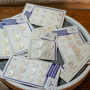 May include: A collection of white plastic buttons on cards with the text "La Mode Buttons" and "Guaranteed Washable".
