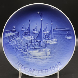Könnte beinhalten: Ein blauer und weißer Weihnachtsteller aus Keramik mit einem Reliefdesign von Fischerbooten, die in einem Hafen segeln. Der Teller ist mit einem Stern und einem Weihnachtsbaum verziert und trägt die Aufschrift "Julgratter 1966".