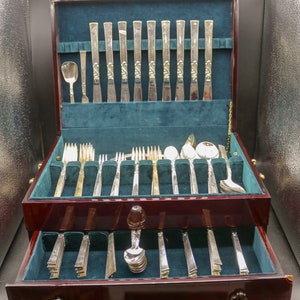 May include: A wooden box with a green velvet interior containing a set of silver cutlery. The cutlery is arranged in two rows, with the top row containing knives, forks, and spoons. The bottom row contains a variety of other utensils, including serving spoons and a butter knife. The box is open and the cutlery is visible.