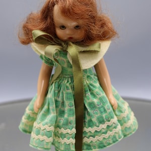 May include: A vintage doll with red hair and a green dress. The dress features a white and green pattern with a matching green ribbon and a white collar. The doll is wearing black shoes.