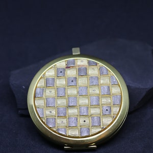 May include: A gold-toned compact mirror with a checkered pattern in silver and gold. The mirror has a round shape and a simple design.