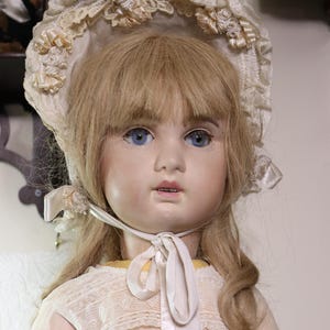 Könnte beinhalten: Eine Porzellanpuppe mit blonden Haaren und blauen Augen, die ein weißes Spitzenhütchen und ein weißes Kleid mit einem Satinband trägt.