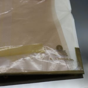 Puede incluir: Una bolsa de pl&aacute;stico transparente parcialmente visible con un interior beige. La bolsa tiene un borde blanco y un sello dorado. El texto "A. E. Mitchell, Inc." es visible en la bolsa.