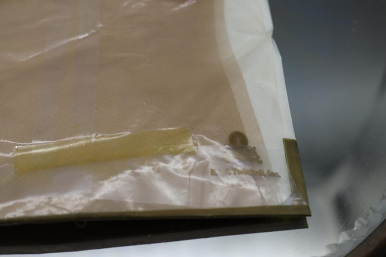Puede incluir: Una bolsa de pl&aacute;stico transparente parcialmente visible con un interior beige. La bolsa tiene un borde blanco y un sello dorado. El texto "A. E. Mitchell, Inc." es visible en la bolsa.