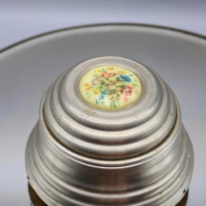 May include: A silver-colored, vintage music box with a floral design on the lid. The lid features a circular, cream-colored insert with a colorful bouquet of flowers. The box has a ribbed, tiered design.