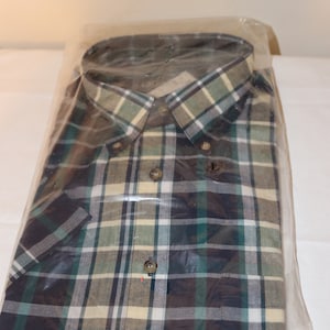 Op de afbeelding: Een geblokt overhemd met knopen in een groene, zwarte en beige kleurstelling. Het overhemd is verpakt in een doorzichtige plastic zak.