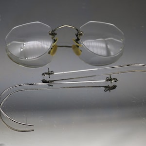 May include: Silver-toned eyeglasses with round frames and a silver-toned wire temple arm. The temple arm is bent into a U-shape.