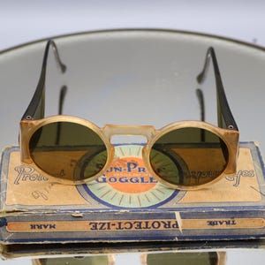 Op de afbeelding: Vintage ronde zonnebril met een lichtbruin montuur en donkere glazen, rustend op een versleten kartonnen doos. De doos is voorzien van de tekst "JN-PR GOGGLE" en "PROTECT-IZE". De zonnebril heeft zwarte pootjes. Een klassiek brillenaccessoire.