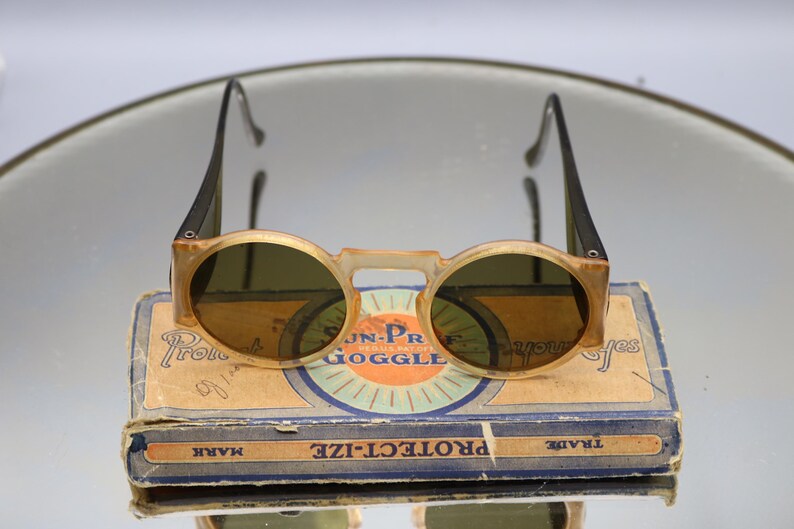 Op de afbeelding: Vintage ronde zonnebril met een lichtbruin montuur en donkere glazen, rustend op een versleten kartonnen doos. De doos is voorzien van de tekst "JN-PR GOGGLE" en "PROTECT-IZE". De zonnebril heeft zwarte pootjes. Een klassiek brillenaccessoire.