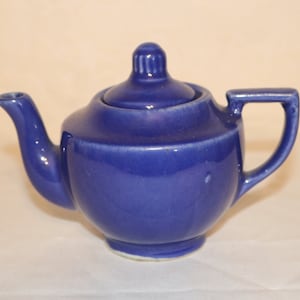 May include: A small, blue ceramic teapot with a lid. The teapot has a rounded body and a spout on the side. The handle is on the opposite side of the spout.