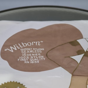 Puede incluir: Un paquete de medias de nailon "Wilborn" Dupont. El paquete presenta un &oacute;valo marr&oacute;n con el nombre de la marca en escritura blanca. El texto dice "SEAMLESS 15 DENIER 100% NYLON FIRST QUALITY RN 18159". La ilustraci&oacute;n muestra piernas de mujer con medias.