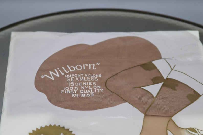 Puede incluir: Un paquete de medias de nailon "Wilborn" Dupont. El paquete presenta un &oacute;valo marr&oacute;n con el nombre de la marca en escritura blanca. El texto dice "SEAMLESS 15 DENIER 100% NYLON FIRST QUALITY RN 18159". La ilustraci&oacute;n muestra piernas de mujer con medias.