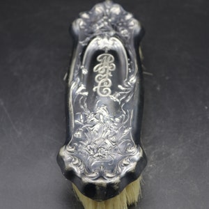 May include: A silver-plated brush with an ornate floral design and the initials "M.C." engraved on the handle. The brush has a natural bristle head.