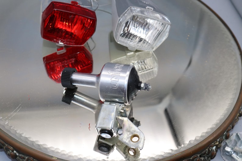 K&ouml;nnte beinhalten: Drei Fahrradlichter, eines rot, eines wei&szlig; und eines silberfarben mit dem Wort "UNION" darauf. Die Lichter befinden sich auf einer spiegelnden Oberfl&auml;che.