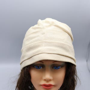 May include: A cream-colored knit beanie hat with a horizontal ribbed design. The hat is displayed on a mannequin head with blue eyes and brown hair. The mannequin has a label that reads "Patty Lehman".