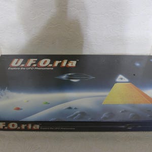 Puede incluir: Una caja rectangular con el título "U.F.O.ria" y el lema "Explore the UFO Phenomena". El arte de la caja presenta una pirámide, un platillo volante y un paisaje con un fondo de cielo nocturno.