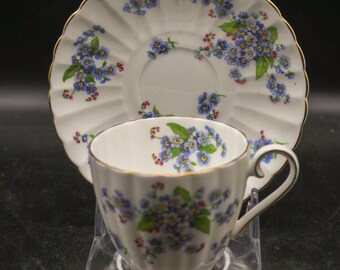 Könnte beinhalten: Eine weiße Teetasse und Untertasse mit einem blauen und rosafarbenen Blumenmuster. Die Untertasse hat einen gewellten Rand und einen goldenen Rand. Die Teetasse hat ein geriffeltes Design.