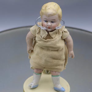 Op de afbeelding: Een kleine porseleinen pop met blond haar, die een beige jurk en blauwe schoenen draagt. De pop staat op een witte sokkel en heeft een zilveren ring om haar nek.