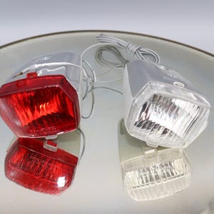K&ouml;nnte beinhalten: Ein Satz mit zwei Fahrradlichtern, eines rot und eines wei&szlig;. Die Lichter sind an einem silbernen Metallrahmen befestigt und haben eine klare Kunststofflinse.