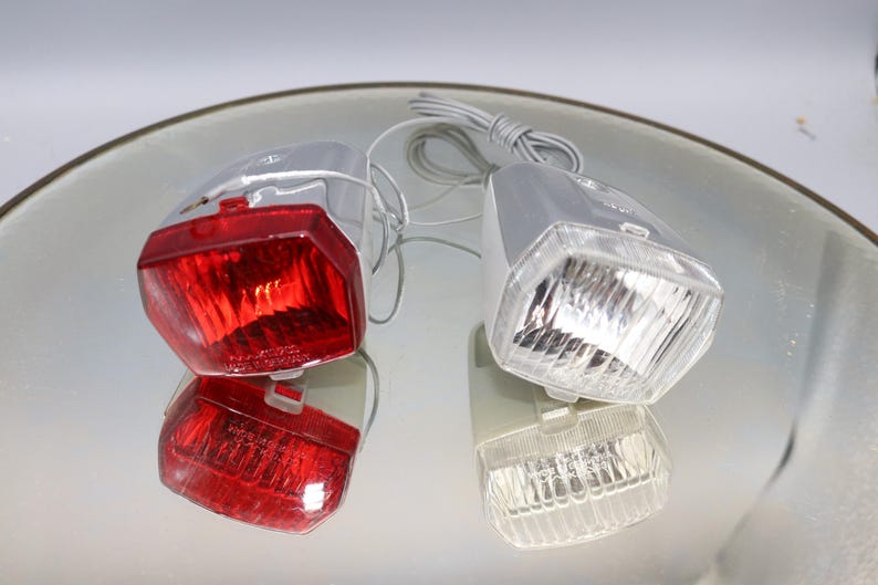 K&ouml;nnte beinhalten: Ein Satz mit zwei Fahrradlichtern, eines rot und eines wei&szlig;. Die Lichter sind an einem silbernen Metallrahmen befestigt und haben eine klare Kunststofflinse.