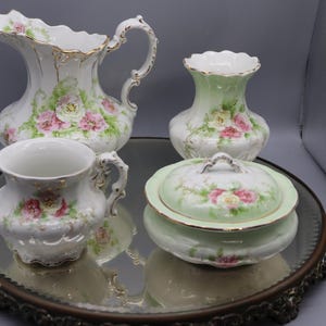 May include: A collection of antique porcelain serving pieces, including a pitcher, vase, cup, and covered dish. Each piece is white with pink rose and green leaf floral designs, accented with gold trim. The set is displayed on a mirrored tray.