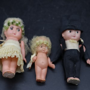 Könnte beinhalten: Drei Vintage-Puppen, eine Braut, ein Bräutigam und eine Blumenmädchen. Die Brautpuppe trägt ein weißes Kleid mit einer Spitzenborte und einer Blumenkrone. Der Bräutigam trägt einen schwarzen Anzug mit einem weißen Hemd und einem schwarzen Zylinder. Das Blumenmädchen trägt ein weißes Kleid mit einer Blumenkrone.