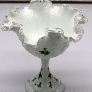 Könnte beinhalten: Eine weiße Milchglas-Bonbonschale mit einem gewellten Rand und einer silbernen Verzierung. Die Schale hat ein Blumenmuster mit grünen Blättern und roten Blüten. Die Schale steht auf einem weißen Sockel.