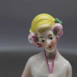 May include: A porcelain figurine of a woman with a yellow hat and pink floral accents. The figurine has a pale complexion, red lips, and a pink ribbon. The figure is a decorative item.