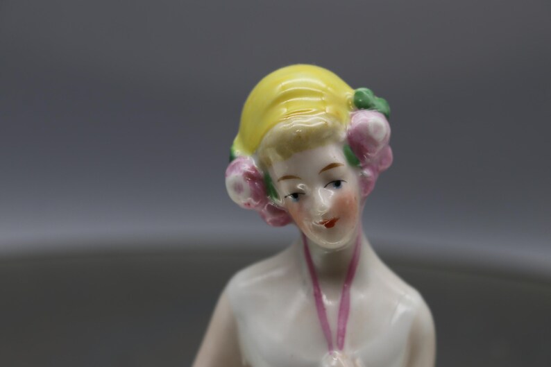 May include: A porcelain figurine of a woman with a yellow hat and pink floral accents. The figurine has a pale complexion, red lips, and a pink ribbon. The figure is a decorative item.