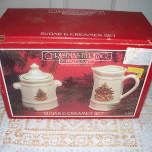Puede incluir: Una caja roja con un juego de azucarero y cremera Christmas Heritage. El juego incluye un azucarero con tapa y una cremera, ambos decorados con un diseño de árbol de Navidad. La caja dice "Sugar & Creamer Set".