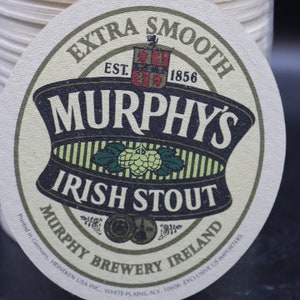 Op de afbeelding: Een ronde, witte bieronderzetter met een groen, zwart en wit ontwerp. De tekst "EXTRA SMOOTH" is gedrukt in een zwart, cursief lettertype bovenaan de onderzetter. De tekst "EST. 1856" is gedrukt in zwart onder de tekst "EXTRA SMOOTH". De tekst "MURPHY'S" is gedrukt in een groot, zwart, cursief lettertype onder de tekst "EST. 1856". De tekst "IRISH STOUT" is gedrukt in een zwart, cursief lettertype onder de tekst "MURPHY'S". De tekst "MURPHY BREWERY IRELAND" is gedrukt in een zwart, cursief lettertype onderaan de onderzetter.