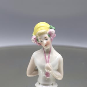 May include: A porcelain half-doll figurine with a yellow hairstyle and pink floral accents. The doll has a white top and a pink necklace. The figurine is a vintage collectible, likely from the early 20th century.