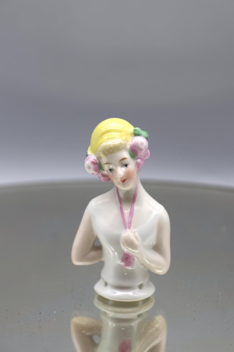 May include: A porcelain half-doll figurine with a yellow hairstyle and pink floral accents. The doll has a white top and a pink necklace. The figurine is a vintage collectible, likely from the early 20th century.