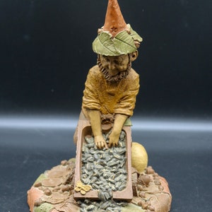 Könnte beinhalten: Eine kleine Gnome-Figur mit einem spitzen Hut und einem Blattwerk. Der Gnome hält eine hölzerne Schubkarre gefüllt mit Sonnenblumenkernen.