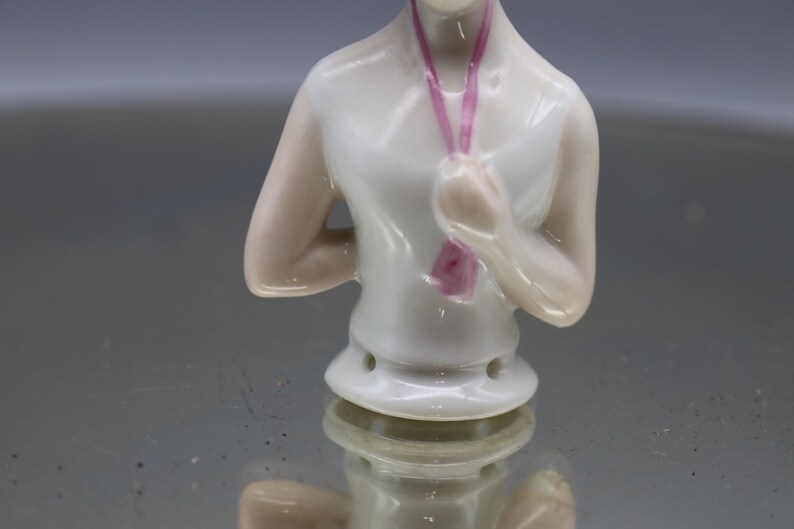 May include: A porcelain doll torso with arms crossed, wearing a white top and a pink ribbon. The doll has a detailed face and is set against a reflective surface. The doll is a vintage collectible.