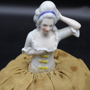 Puede incluir: Una muñeca de porcelana en forma de alfiletero, vestida con un vestido blanco y una cinta azul en el pelo. Lleva un corsé amarillo y blanco y una falda de tela marrón con lunares blancos.
